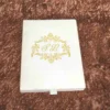A high-angle, close-up shot of a **luxurious invitation box** with an ornate, three-dimensional lid design. The box features: * A **laser-cut gold wooden frame** on the lid with intricate floral and scrollwork patterns. * A central ** plaque** with a custom gold-etched monogram "R & F" in an elegant script. * A border of **sparkling clear crystals** embedded along the inner edge of the gold frame, adding a high-shine detail. * The rich, deep red velvet fabric covers the sides of the box, providing a soft, tactile contrast to the metallic gold top. * The box is displayed on a neutral, textured light-colored surface, highlighting its sophisticated and artisanal craftsmanship.