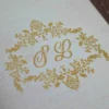 A high-angle, close-up shot of a **luxurious invitation box** with an ornate, three-dimensional lid design. The box features: * A **laser-cut gold wooden frame** on the lid with intricate floral and scrollwork patterns. * A central ** plaque** with a custom gold-etched monogram "R & F" in an elegant script. * A border of **sparkling clear crystals** embedded along the inner edge of the gold frame, adding a high-shine detail. * The rich, deep red velvet fabric covers the sides of the box, providing a soft, tactile contrast to the metallic gold top. * The box is displayed on a neutral, textured light-colored surface, highlighting its sophisticated and artisanal craftsmanship.