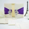 An open, ivory-colored fabric gatefold invitation box displaying a formal baptism announcement. The main invitation card is nestled in the center panel, featuring elegant silver scrollwork at the top corners and script text for the name "Reese Ava Jeanne Sloan." The two side flaps of the box include pockets. In the foreground, several matching smaller cards, including an RSVP and an Information card, are laid out on a white surface. The purple ribbon and crystal brooch from the exterior are visible on the outer edges of the open flaps.