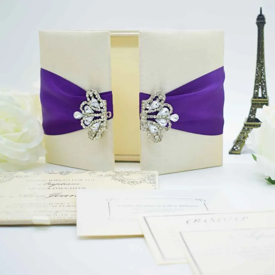 An open, ivory-colored fabric gatefold invitation box displaying a formal baptism announcement. The main invitation card is nestled in the center panel, featuring elegant silver scrollwork at the top corners and script text for the name "Reese Ava Jeanne Sloan." The two side flaps of the box include pockets. In the foreground, several matching smaller cards, including an RSVP and an Information card, are laid out on a white surface. The purple ribbon and crystal brooch from the exterior are visible on the outer edges of the open flaps.