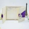 An open, ivory-colored fabric gatefold invitation box displaying a formal baptism announcement. The main invitation card is nestled in the center panel, featuring elegant silver scrollwork at the top corners and script text for the name "Reese Ava Jeanne Sloan." The two side flaps of the box include pockets. In the foreground, several matching smaller cards, including an RSVP and an Information card, are laid out on a white surface. The purple ribbon and crystal brooch from the exterior are visible on the outer edges of the open flaps.