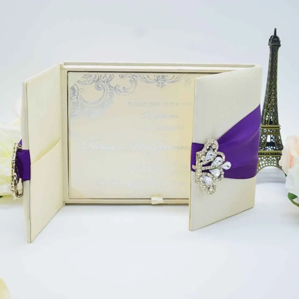 An open, ivory-colored fabric gatefold invitation box displaying a formal baptism announcement. The main invitation card is nestled in the center panel, featuring elegant silver scrollwork at the top corners and script text for the name "Reese Ava Jeanne Sloan." The two side flaps of the box include pockets. In the foreground, several matching smaller cards, including an RSVP and an Information card, are laid out on a white surface. The purple ribbon and crystal brooch from the exterior are visible on the outer edges of the open flaps.