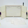 An open, ivory-colored fabric gatefold invitation box displaying a formal baptism announcement. The main invitation card is nestled in the center panel, featuring elegant silver scrollwork at the top corners and script text for the name "Reese Ava Jeanne Sloan." The two side flaps of the box include pockets. In the foreground, several matching smaller cards, including an RSVP and an Information card, are laid out on a white surface. The purple ribbon and crystal brooch from the exterior are visible on the outer edges of the open flaps.
