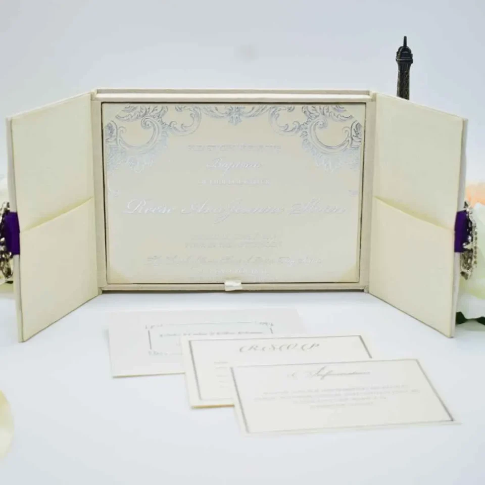 An open, ivory-colored fabric gatefold invitation box displaying a formal baptism announcement. The main invitation card is nestled in the center panel, featuring elegant silver scrollwork at the top corners and script text for the name "Reese Ava Jeanne Sloan." The two side flaps of the box include pockets. In the foreground, several matching smaller cards, including an RSVP and an Information card, are laid out on a white surface. The purple ribbon and crystal brooch from the exterior are visible on the outer edges of the open flaps.