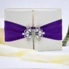 An open, ivory-colored fabric gatefold invitation box displaying a formal baptism announcement. The main invitation card is nestled in the center panel, featuring elegant silver scrollwork at the top corners and script text for the name "Reese Ava Jeanne Sloan." The two side flaps of the box include pockets. In the foreground, several matching smaller cards, including an RSVP and an Information card, are laid out on a white surface. The purple ribbon and crystal brooch from the exterior are visible on the outer edges of the open flaps.