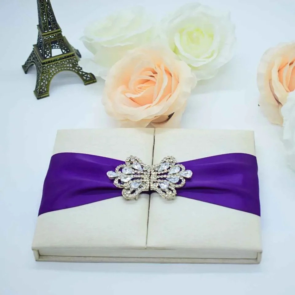 An open, ivory-colored fabric gatefold invitation box displaying a formal baptism announcement. The main invitation card is nestled in the center panel, featuring elegant silver scrollwork at the top corners and script text for the name "Reese Ava Jeanne Sloan." The two side flaps of the box include pockets. In the foreground, several matching smaller cards, including an RSVP and an Information card, are laid out on a white surface. The purple ribbon and crystal brooch from the exterior are visible on the outer edges of the open flaps.