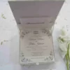 An open, square ivory silk invitation box shown from a high angle. The lid is propped open, revealing a pocket containing a small card with the names "Oliver and Emily" in script. The main wedding invitation is nestled inside the box, featuring black ornate scrollwork at the corners and elegant typography for the names "Victoria Marie and Patric Johnson." The box sits on a white surface scattered with light-colored dried petals, with a white flower stem visible on the right.