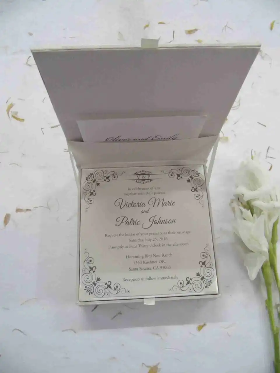 An open, square ivory silk invitation box shown from a high angle. The lid is propped open, revealing a pocket containing a small card with the names "Oliver and Emily" in script. The main wedding invitation is nestled inside the box, featuring black ornate scrollwork at the corners and elegant typography for the names "Victoria Marie and Patric Johnson." The box sits on a white surface scattered with light-colored dried petals, with a white flower stem visible on the right.