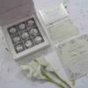 An open, ivory silk favor box featuring a nine-compartment grid, with each section holding a chocolate truffle wrapped in textured silver foil. To the right of the box, the matching wedding stationery suite is displayed, including an RSVP card and a formal invitation for Victoria Marie and Patric Johnson decorated with elegant black scrollwork. Two white gladiolus blooms and scattered dried petals rest on the light-colored surface in the foreground.