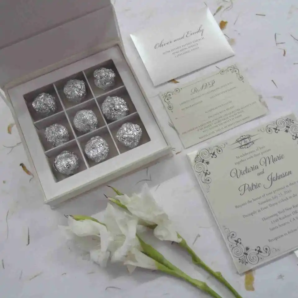 An open, ivory silk favor box featuring a nine-compartment grid, with each section holding a chocolate truffle wrapped in textured silver foil. To the right of the box, the matching wedding stationery suite is displayed, including an RSVP card and a formal invitation for Victoria Marie and Patric Johnson decorated with elegant black scrollwork. Two white gladiolus blooms and scattered dried petals rest on the light-colored surface in the foreground.
