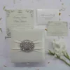 A top-down view of a square, ivory silk invitation box. A matching ivory satin ribbon is wrapped horizontally across the center, gathered in the middle by a large, oval-shaped crystal brooch that features iridescent, multicolored rhinestones. The box sits on a white surface scattered with delicate, dried flower petals, with a sprig of white gladiolus flowers resting on the right side.