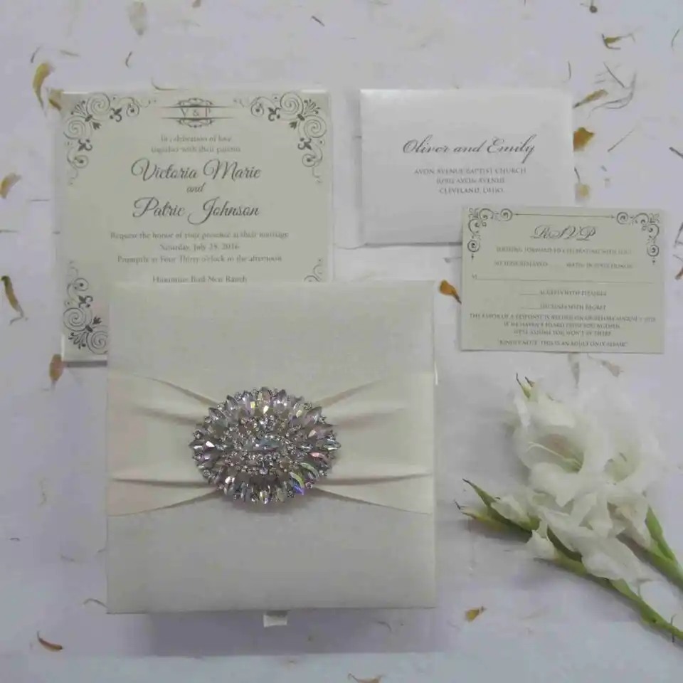 A top-down view of a square, ivory silk invitation box. A matching ivory satin ribbon is wrapped horizontally across the center, gathered in the middle by a large, oval-shaped crystal brooch that features iridescent, multicolored rhinestones. The box sits on a white surface scattered with delicate, dried flower petals, with a sprig of white gladiolus flowers resting on the right side.