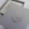 A top-down view of a square, ivory silk invitation box. A matching ivory satin ribbon is wrapped horizontally across the center, gathered in the middle by a large, oval-shaped crystal brooch that features iridescent, multicolored rhinestones. The box sits on a white surface scattered with delicate, dried flower petals, with a sprig of white gladiolus flowers resting on the right side.