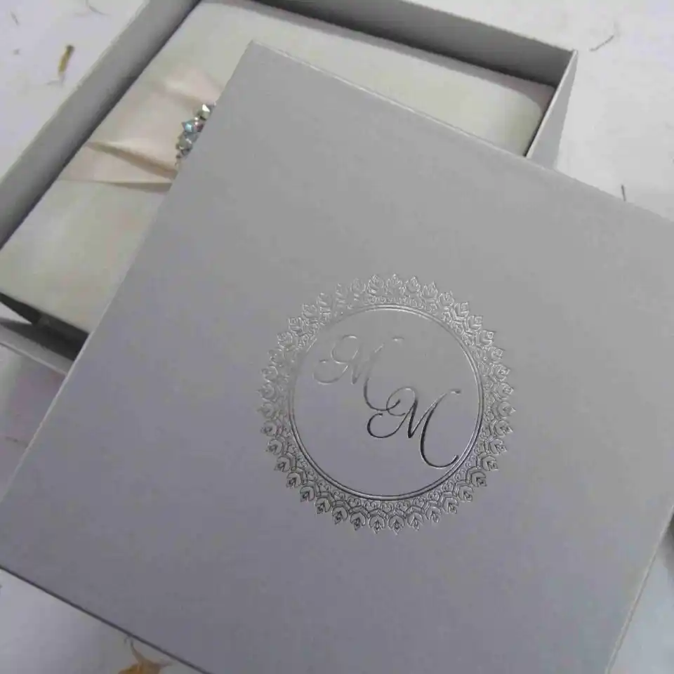 A top-down view of a square, ivory silk invitation box. A matching ivory satin ribbon is wrapped horizontally across the center, gathered in the middle by a large, oval-shaped crystal brooch that features iridescent, multicolored rhinestones. The box sits on a white surface scattered with delicate, dried flower petals, with a sprig of white gladiolus flowers resting on the right side.