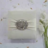 A top-down view of a square, ivory silk invitation box. A matching ivory satin ribbon is wrapped horizontally across the center, gathered in the middle by a large, oval-shaped crystal brooch that features iridescent, multicolored rhinestones. The box sits on a white surface scattered with delicate, dried flower petals, with a sprig of white gladiolus flowers resting on the right side.