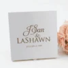 A top-down, slightly angled shot of a **square white fabric wedding invitation box** featuring elegant metallic detailing. The box is characterized by: * **Gold or bronze foil lettering** centered on the lid, reading "J'San & LASHAWN" in a mix of script and serif fonts. * The wedding date, "JANUARY 20, 2019," printed in smaller matching font below the names. * A small **white satin pull-tab** visible at the bottom center of the lid. * A cluster of **pale peach roses** positioned in the upper right corner, partially framing the box. * A clean, minimalist white background that highlights the crisp texture of the fabric and the shimmer of the foil.