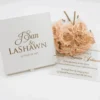 A high-angle, slightly tilted shot of a **white fabric invitation box** and a clear **acrylic invitation card** from two different event suites, arranged on a minimalist white surface. The composition includes: * **The Box (Foreground):** A square white fabric box featuring the names "J'San & LASHAWN" and the date "JANUARY 20, 2019" embossed in elegant gold foil. A small white satin pull-tab is visible at the bottom. * **The Acrylic Card (Background):** A clear acrylic invitation for "Queen Annette Johnson" featuring gold-foiled typography and a large royal crest with the monogram "AJ." * **Accents:** A bouquet of **pale peach roses** rests between the two items, adding a soft, romantic touch to the presentation. * The bright lighting emphasizes the reflective gold foil and the crisp, clean textures of the stationery.