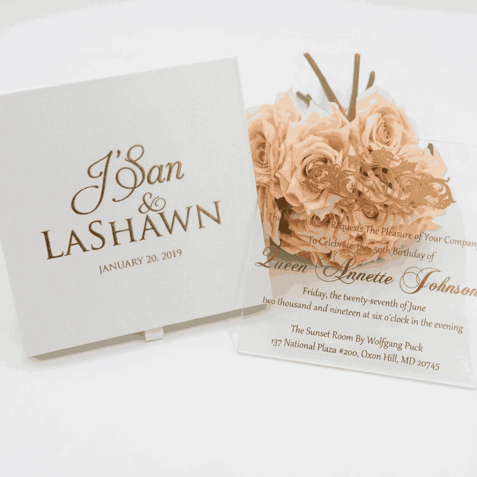 A high-angle, slightly tilted shot of a **white fabric invitation box** and a clear **acrylic invitation card** from two different event suites, arranged on a minimalist white surface. The composition includes: * **The Box (Foreground):** A square white fabric box featuring the names "J'San & LASHAWN" and the date "JANUARY 20, 2019" embossed in elegant gold foil. A small white satin pull-tab is visible at the bottom. * **The Acrylic Card (Background):** A clear acrylic invitation for "Queen Annette Johnson" featuring gold-foiled typography and a large royal crest with the monogram "AJ." * **Accents:** A bouquet of **pale peach roses** rests between the two items, adding a soft, romantic touch to the presentation. * The bright lighting emphasizes the reflective gold foil and the crisp, clean textures of the stationery.