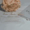 A high-angle, slightly tilted shot of a **white fabric invitation box** and a clear **acrylic invitation card** from two different event suites, arranged on a minimalist white surface. The composition includes: * **The Box (Foreground):** A square white fabric box featuring the names "J'San & LASHAWN" and the date "JANUARY 20, 2019" embossed in elegant gold foil. A small white satin pull-tab is visible at the bottom. * **The Acrylic Card (Background):** A clear acrylic invitation for "Queen Annette Johnson" featuring gold-foiled typography and a large royal crest with the monogram "AJ." * **Accents:** A bouquet of **pale peach roses** rests between the two items, adding a soft, romantic touch to the presentation. * The bright lighting emphasizes the reflective gold foil and the crisp, clean textures of the stationery.