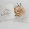 A high-angle, slightly tilted shot of a **white fabric invitation box** and a clear **acrylic invitation card** from two different event suites, arranged on a minimalist white surface. The composition includes: * **The Box (Foreground):** A square white fabric box featuring the names "J'San & LASHAWN" and the date "JANUARY 20, 2019" embossed in elegant gold foil. A small white satin pull-tab is visible at the bottom. * **The Acrylic Card (Background):** A clear acrylic invitation for "Queen Annette Johnson" featuring gold-foiled typography and a large royal crest with the monogram "AJ." * **Accents:** A bouquet of **pale peach roses** rests between the two items, adding a soft, romantic touch to the presentation. * The bright lighting emphasizes the reflective gold foil and the crisp, clean textures of the stationery.