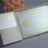 An open, ivory silk gatefold invitation box revealing a formal wedding invitation for Mia Dimitrijevic and David Baker. The invitation features elegant gold script and a gold-stamped circular monogram on the left side. The card is framed with a thin gold border. The left flap of the box contains a pocket holding a matching RSVP card. A bouquet of soft peach-colored roses sits in the background on a light grey surface.