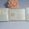 An open, ivory silk gatefold invitation box revealing a formal wedding invitation for Mia Dimitrijevic and David Baker. The invitation features elegant gold script and a gold-stamped circular monogram on the left side. The card is framed with a thin gold border. The left flap of the box contains a pocket holding a matching RSVP card. A bouquet of soft peach-colored roses sits in the background on a light grey surface.