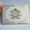 A luxurious, ivory silk gatefold invitation box featuring elaborate gold foil accents. The four corners are decorated with intricate, swirling baroque-style patterns. In the center, a large, ornate gold crest features a crown at the top and a circular monogram with the letters "MY" in the middle. Below the monogram, a gold banner displays the date "26-11-2015." The invitation sits on a light grey surface with a soft-focus bouquet of peach roses visible in the upper left corner.