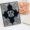 A sophisticated, black rectangular invitation card featuring striking silver-embossed details. Highly decorative, baroque-style silver scrollwork accents each of the four corners. In the center, an interlocking silver monogram stands out against the dark background. The card is displayed upright on a white surface next to a bouquet of soft peach-colored roses and a closed white notebook.