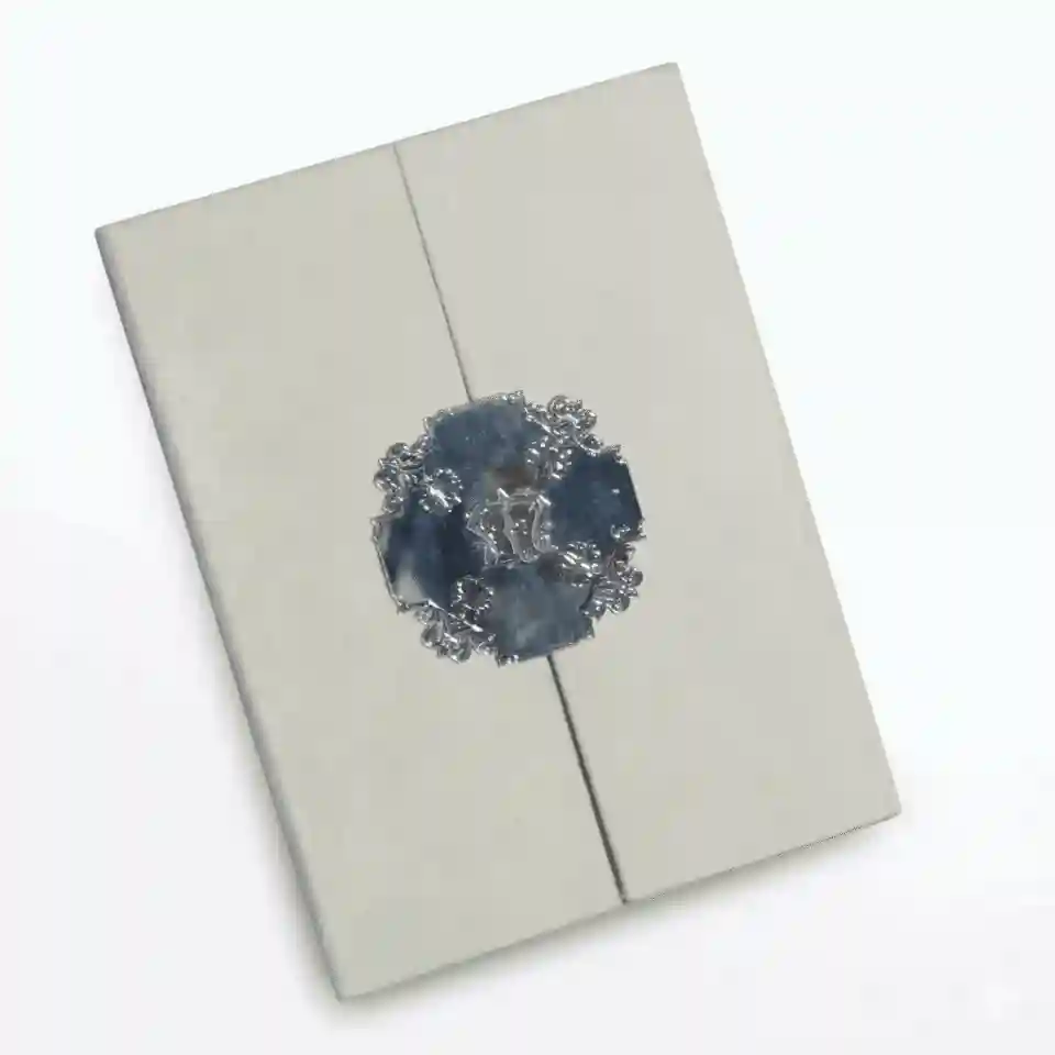 An open, ivory silk invitation box shown against a plain white background. The interior lid features a laser-cut silver filigree pocket holding a cream-colored card with silver foil accents. The bottom of the box contains a padded insert with a silver mirror-finish acrylic monogram plate in the center, featuring an ornate crest. The box is shown from a high-angle perspective, highlighting its deep, luxurious structure.