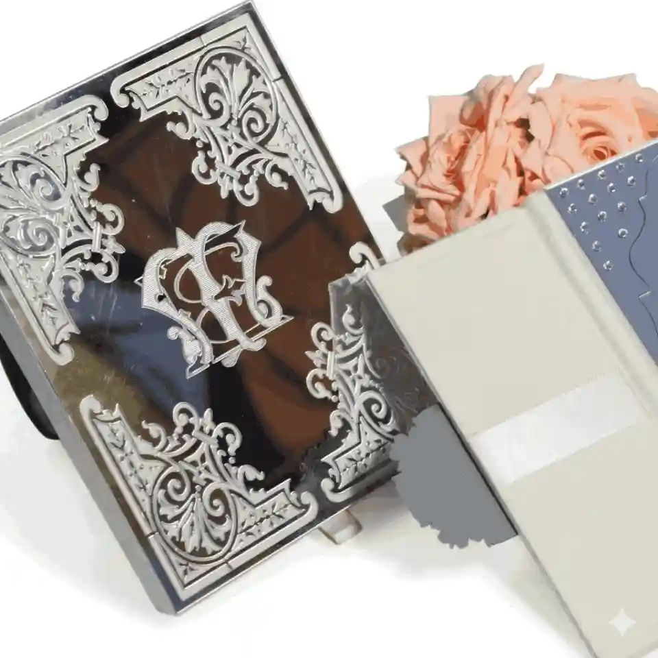 A sophisticated, black rectangular invitation card featuring striking silver-embossed details. Highly decorative, baroque-style silver scrollwork accents each of the four corners. In the center, an interlocking silver monogram stands out against the dark background. The card is displayed upright on a white surface next to a bouquet of soft peach-colored roses and a closed white notebook.