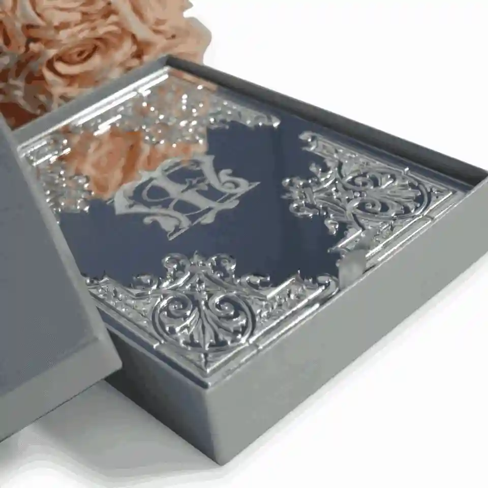 A sophisticated, black rectangular invitation card featuring striking silver-embossed details. Highly decorative, baroque-style silver scrollwork accents each of the four corners. In the center, an interlocking silver monogram stands out against the dark background. The card is displayed upright on a white surface next to a bouquet of soft peach-colored roses and a closed white notebook.