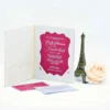 An open, off-white velvet folio wedding invitation is displayed on a white surface, accompanied by decorative elements. * **The Folio:** The left interior panel features an empty scalloped pocket. The right panel holds the main invitation card with white satin corner ribbons. * **The Invitation Card:** A magenta, bracket-edged die-cut card is layered over a shimmering silver glitter background. It features white script announcing the wedding of **Emily Echevarria and Brandon Hirsch**. * **Inserts:** Resting on the surface in front of the folio are a matching magenta RSVP card and a small white envelope. * **Decor:** To the right stands a bronze-toned Eiffel Tower figurine and a soft, peach-colored rose.