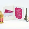 An open, off-white velvet folio wedding invitation is displayed against a white background. * **Left Side:** A scalloped pocket contains a magenta RSVP card with white script and a matching envelope featuring the names "Emily and Brandon." * **Right Side:** The main invitation card is held by white corner ribbons. It features a magenta, bracket-edged frame containing white text that announces the marriage of **Emily Echevarria and Brandon Hirsch**. The card is layered over a shimmering silver glitter backing. The scene is decorated with a gold lantern containing a magenta candle on the left, and a bronze Eiffel Tower figurine with a pale rose on the right.