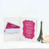 An open, off-white velvet folio wedding invitation is displayed against a white background. * **Left Side:** A scalloped pocket contains a magenta RSVP card with white script and a matching envelope featuring the names "Emily and Brandon." * **Right Side:** The main invitation card is held by white corner ribbons. It features a magenta, bracket-edged frame containing white text that announces the marriage of **Emily Echevarria and Brandon Hirsch**. The card is layered over a shimmering silver glitter backing. The scene is decorated with a gold lantern containing a magenta candle on the left, and a bronze Eiffel Tower figurine with a pale rose on the right.