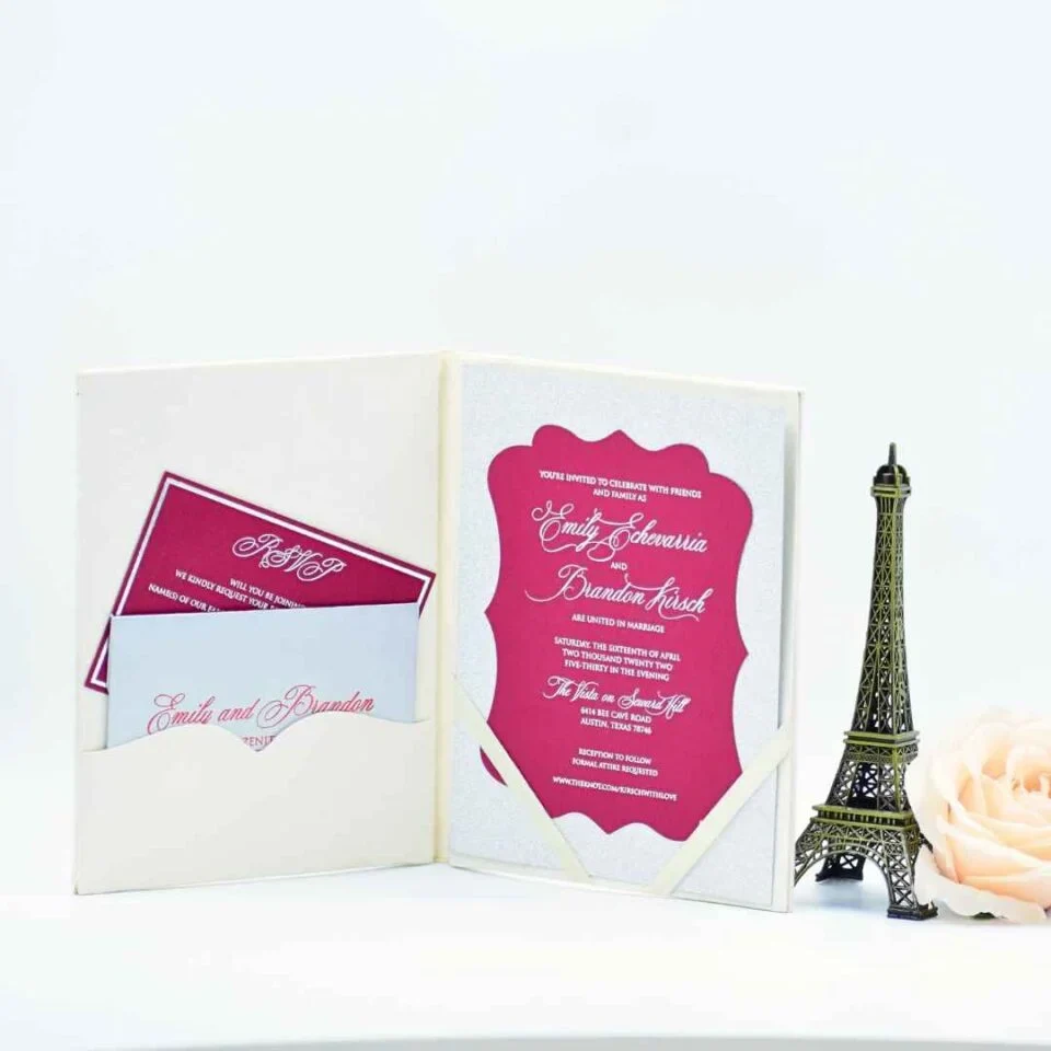 An open, off-white velvet folio wedding invitation is displayed against a white background. * **Left Side:** A scalloped pocket contains a magenta RSVP card with white script and a matching envelope featuring the names "Emily and Brandon." * **Right Side:** The main invitation card is held by white corner ribbons. It features a magenta, bracket-edged frame containing white text that announces the marriage of **Emily Echevarria and Brandon Hirsch**. The card is layered over a shimmering silver glitter backing. The scene is decorated with a gold lantern containing a magenta candle on the left, and a bronze Eiffel Tower figurine with a pale rose on the right.