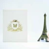 A portrait-oriented, off-white velvet invitation folder stands upright against a clean white background. Centered on the cover is a detailed gold foil crest featuring a crown, floral embellishments, and a central monogram. A gold ribbon banner at the bottom of the crest reads with the date To the right of the invitation, a small bronze-colored Eiffel Tower figurine and a hint of a pale rose add a decorative touch.