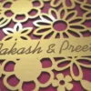 A high-angle shot of a square, chocolate-brown invitation box. The lid features an intricate, laser-cut gold floral pattern of various daisies and blossoms over a vibrant magenta background. A gold banner integrated into the floral design is engraved with the names "Aakash & Preeti" in an elegant script. A second, identical box is partially visible in the blurred background.