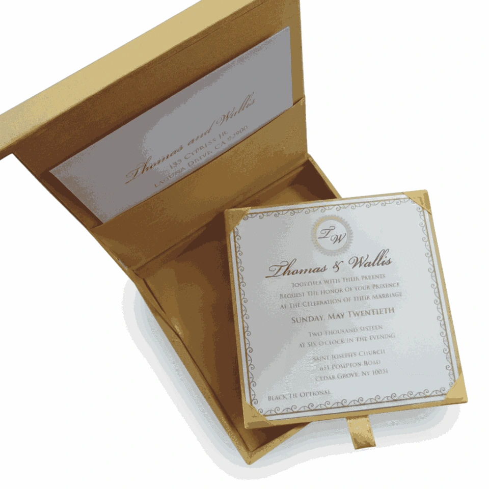 An open, square gold silk invitation box shown from an angled top-down perspective against a white background. The interior lid features a pocket holding a card with the names "Thomas and Wallis" in gold script. The main wedding invitation is held in the base of the box by four gold corner ribbons. The invitation card displays a circular "TW" monogram at the top, elegant typography for the wedding details of Thomas and Wallis, and a decorative silver scrollwork border. A small gold silk pull-tab is visible at the bottom edge of the box.