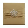 A top-down view of a square, gold silk invitation folio isolated against a white background. A matching gold satin ribbon is wrapped horizontally across the center, cinched by a large, circular brooch featuring white pearls and small, sparkling rhinestones. The fabric has a subtle, elegant sheen.