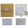 An open, square gold silk invitation box shown from an angled top-down perspective against a white background. The interior lid features a pocket holding a card with the names "Thomas and Wallis" in gold script. The main wedding invitation is held in the base of the box by four gold corner ribbons. The invitation card displays a circular "TW" monogram at the top, elegant typography for the wedding details of Thomas and Wallis, and a decorative silver scrollwork border. A small gold silk pull-tab is visible at the bottom edge of the box.