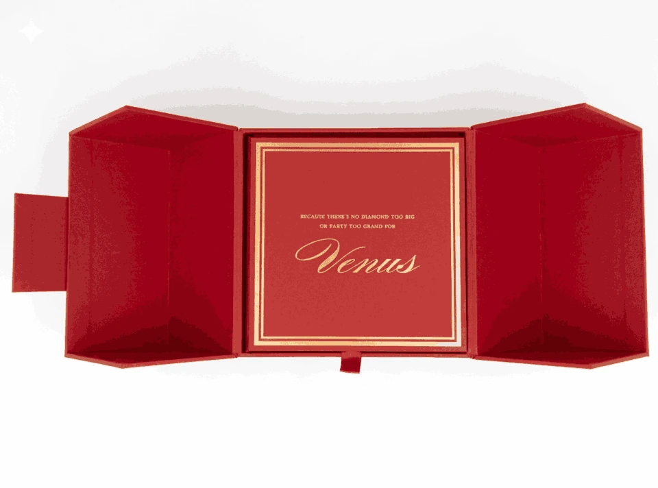 An overhead view of the open, deep red invitation box from the previous image. The two side flaps are spread wide, showing their angled, geometric construction. Inside the central compartment, a matching red card is framed by a double gold border. The card features the word "Venus" in elegant gold script, with a smaller line of gold text above it that reads, "Because there's no diamond too big or party too grand for." A small red ribbon pull-tab is visible at the bottom center of the box.