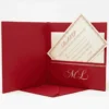 An open, deep red invitation box featuring a padded interior. Inside the lid, a matching red pocket holds a cream-colored card with the names "Oliver and Emily" and an address in Cleveland, Ohio, printed in elegant gold script. A gold-stamped "ML" monogram sits just below the pocket. The base of the box contains a white invitation card with a decorative gold border. The entire ensemble is set against a clean, white background.