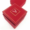 A high-angle shot of a deep red, square invitation box with a hinged lid. The top of the box is decorated with a thin, ornate gold foil border. In the center, a gold-framed square panel features a stylized letter "V" created from small, sparkling clear crystals. The box is shown slightly open, and its matching red lid sits separately in the background on a clean white surface.