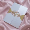 A close-up shot shows a gold, laser-cut paper belly band securing a light blue or silver shimmering gatefold invitation. The focal point is an intricate, symmetrical monogram or Celtic-style knot design in the center, featuring elegant swirls and clean geometric lines. The gold paper has a subtle metallic texture that contrasts with the fine-grained, pearlescent finish of the invitation cardstock underneath. The vertical seam where the two panels of the gatefold meet is visible directly behind the center of the gold ornament, highlighting the precision of the packaging.