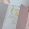 A close-up shot shows a gold, laser-cut paper belly band securing a light blue or silver shimmering gatefold invitation. The focal point is an intricate, symmetrical monogram or Celtic-style knot design in the center, featuring elegant swirls and clean geometric lines. The gold paper has a subtle metallic texture that contrasts with the fine-grained, pearlescent finish of the invitation cardstock underneath. The vertical seam where the two panels of the gatefold meet is visible directly behind the center of the gold ornament, highlighting the precision of the packaging.