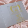 A close-up shot shows a gold, laser-cut paper belly band securing a light blue or silver shimmering gatefold invitation. The focal point is an intricate, symmetrical monogram or Celtic-style knot design in the center, featuring elegant swirls and clean geometric lines. The gold paper has a subtle metallic texture that contrasts with the fine-grained, pearlescent finish of the invitation cardstock underneath. The vertical seam where the two panels of the gatefold meet is visible directly behind the center of the gold ornament, highlighting the precision of the packaging.