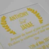 A close-up shot shows a gold, laser-cut paper belly band securing a light blue or silver shimmering gatefold invitation. The focal point is an intricate, symmetrical monogram or Celtic-style knot design in the center, featuring elegant swirls and clean geometric lines. The gold paper has a subtle metallic texture that contrasts with the fine-grained, pearlescent finish of the invitation cardstock underneath. The vertical seam where the two panels of the gatefold meet is visible directly behind the center of the gold ornament, highlighting the precision of the packaging.