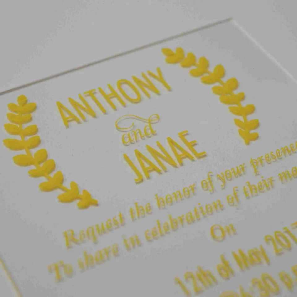 A close-up shot shows a gold, laser-cut paper belly band securing a light blue or silver shimmering gatefold invitation. The focal point is an intricate, symmetrical monogram or Celtic-style knot design in the center, featuring elegant swirls and clean geometric lines. The gold paper has a subtle metallic texture that contrasts with the fine-grained, pearlescent finish of the invitation cardstock underneath. The vertical seam where the two panels of the gatefold meet is visible directly behind the center of the gold ornament, highlighting the precision of the packaging.