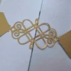 A close-up shot shows a gold, laser-cut paper belly band securing a light blue or silver shimmering gatefold invitation. The focal point is an intricate, symmetrical monogram or Celtic-style knot design in the center, featuring elegant swirls and clean geometric lines. The gold paper has a subtle metallic texture that contrasts with the fine-grained, pearlescent finish of the invitation cardstock underneath. The vertical seam where the two panels of the gatefold meet is visible directly behind the center of the gold ornament, highlighting the precision of the packaging.