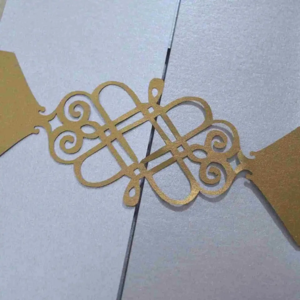 A close-up shot shows a gold, laser-cut paper belly band securing a light blue or silver shimmering gatefold invitation. The focal point is an intricate, symmetrical monogram or Celtic-style knot design in the center, featuring elegant swirls and clean geometric lines. The gold paper has a subtle metallic texture that contrasts with the fine-grained, pearlescent finish of the invitation cardstock underneath. The vertical seam where the two panels of the gatefold meet is visible directly behind the center of the gold ornament, highlighting the precision of the packaging.
